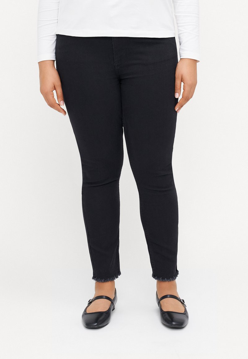 Jeans negros ajustados con dobladillos deshilachados, hechos de denim; combinados con bailarinas negras que tienen una correa sobre el empeine. También se ve una blusa blanca de mangas largas.