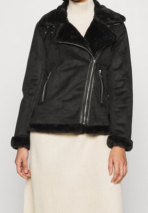 Schwarze Bikerjacke mit Kunstpelz-Futter, asymmetrischem silbernem Reißverschluss, seitlichen Reißverschlusstaschen, getragen über einem cremefarbenen Strickkleid.