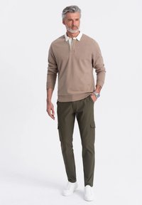 Beige sweatshirt med vit krage, ihop med olivgröna cargobyxor. Modellen bär vita sneakers och ett silverfärgat armbandsur. Texturerat tyg.