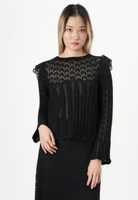 Kvinne med langt hår iført en svart blond strikket langermet topp og matchende svart blond skjørt, stående mot en hvit bakgrunn.