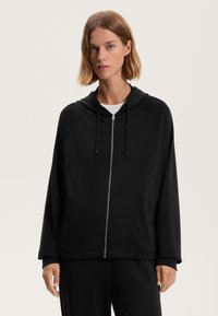 Črna zadrgo zapeta kapuca s kapuco, raglan rokavi in stranskimi žepi. Narejena iz mehkega blaga z gladko teksturo in sproščenim krojem.