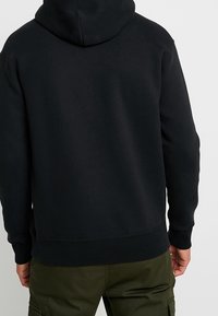 Sudadera negra con capucha de textura suave, puños y dobladillo acanalados. Presenta un pequeño logotipo en la parte posterior, cerca del área del cuello. El usuario lleva pantalones verdes.