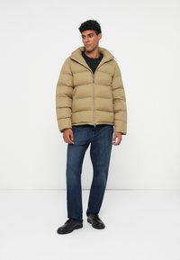 Beige puffatakki, jossa on vetoketjullinen etuosa, suuri huppu ja teksturoitu ommel. Yhdistetty tummansinsisiin farkkuihin ja mustiin kenkiin.