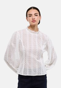 Blouse blanche avec broderie dentelle, col haut, manches bouffantes et motifs découpés complexes sur toute la surface.