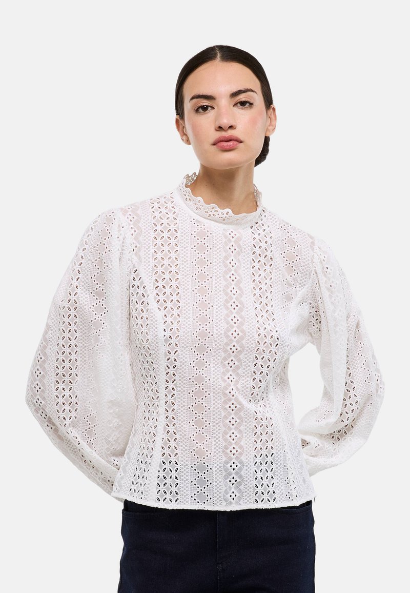 Blouse blanche avec broderie dentelle, col haut, manches bouffantes et motifs découpés complexes sur toute la surface.