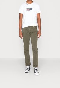 Biała koszulka z granatowym logo i czerwonymi paskami na klatce piersiowej, w połączeniu z oliwkowymi spodniami i czarnymi wysokimi sneakersami.