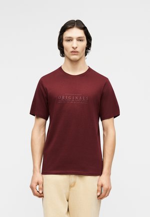 Νεαρός άνδρας με σκουρόχρωμα μαλλιά φοράει μπορντό μπλούζα με το κείμενο «Originals» και μπεζ παντελόνι, στέκεται μπροστά σε ένα απλό φωτεινό φόντο.