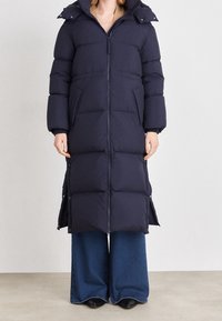 Marineblauer langer Steppmantel mit Reißverschluss vorne, Kapuze und Seitentaschen. Gepolstertes Design mit glatter Textur. Kombiniert mit weit geschnittenen Jeans.