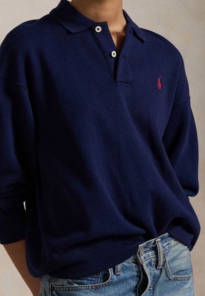 Isik kannab tumesinist pika varrukaga polosärki, millel on kaks nuppu ja väike punane logo, koos siniste teksadega.