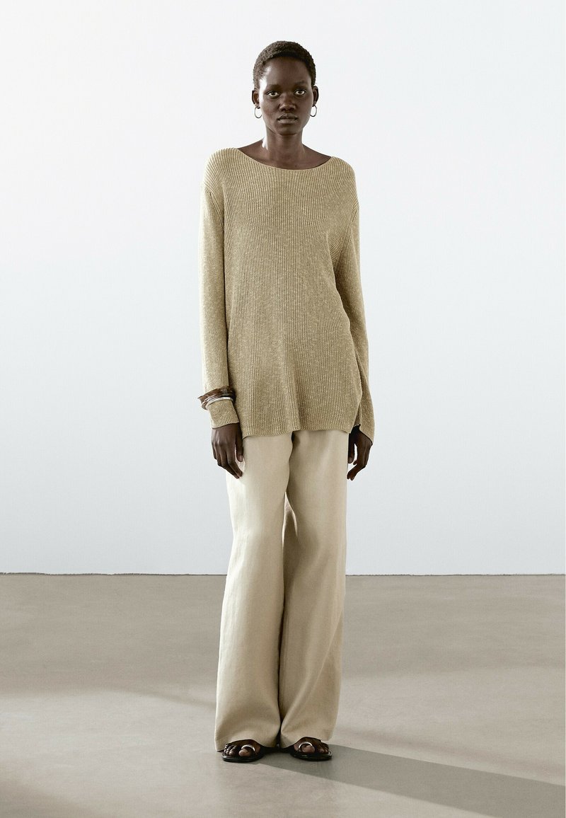 Maglione a coste di un beige chiaro con scollatura a barca, abbinato a pantaloni larghi di crema e sandali scuri, creando una silhouette rilassata.