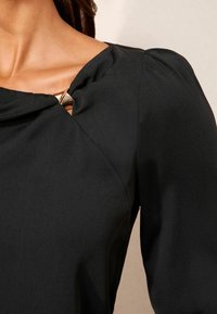 Bluza neagră cu decolteu drapat și un accent metalic la umăr. Materialul are o textură netedă și un luciu ușor.