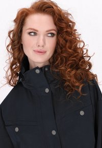 Marineblauwe waterdichte jas met hoge kraag, drukknoopsluiting en twee borstzakken. Beschikt over een ontspannen pasvorm en een gladde stoftextuur.