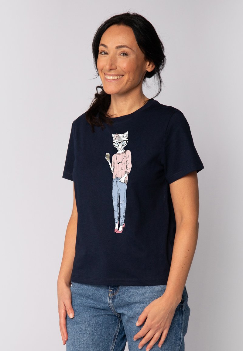 Navy blauwe katoenen t-shirt met een grafisch ontwerp van een kat met een bril en een trui, gecombineerd met lichtblauwe jeans en roze schoenen.