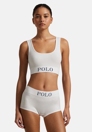 Vrouw draagt een witte geribbelde cropped top en short set met het "POLO" logo op de tailleband, staand tegen een effen lichte achtergrond.