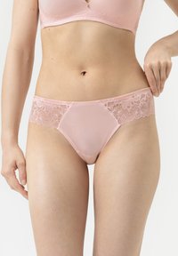 Femme portant un soutien-gorge en dentelle rose clair et un slip assorti, ajustant la taille sur le côté droit, montrée de la poitrine jusqu'à la partie supérieure des cuisses.