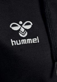 Logo hummel brodé blanc avec bourdon stylisé et texte sur tissu noir, à côté d'un cordon tressé noir.