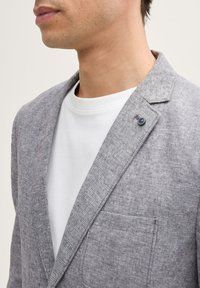 Grauer Blazer aus strukturiertem Stoff mit Reverskragen und Brusttasche. Trägt er über einem weißen Rundhals-T-Shirt mit einem kleinen Knopf-Detail.