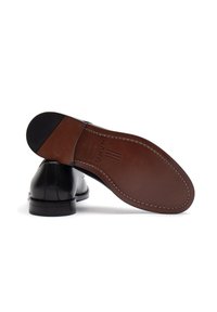 Scarpe da uomo in pelle nera con finitura liscia, dotate di una suola marrone con cuciture e un tacco strutturato. Nome del brand impresso sulla suola.