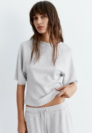 Giovane donna con lunghi capelli castani che indossa una maglietta a maniche corte grigio chiaro e pantaloni della tuta abbinati, sollevando l'orlo della maglietta per mostrare l'addome.