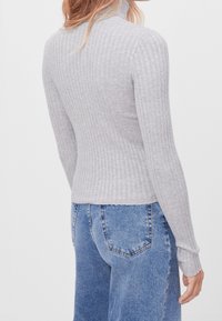 Femme portant un pull à col roulé côtelé gris clair et un jean en denim bleu, vue de dos sur fond blanc uni.