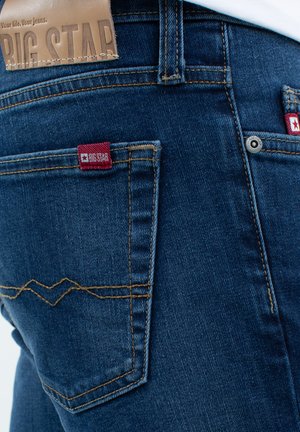 Mėlynos džinsinės kelnes su šiek tiek išblukusia tekstūra; turi raudoną logotipo etiketę ant užpakalinio kišenės ir dekoratyvinį siuvimą zigzagu.