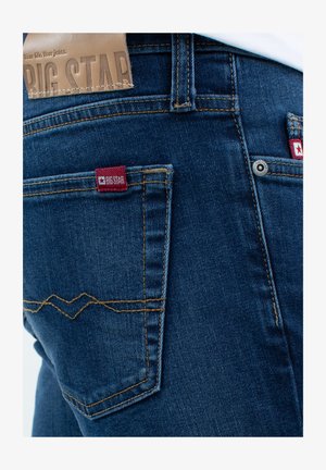 Blaue Denim-Jeans mit einer leicht ausgebleichten Textur; verfügen über ein rotes Logoetikett an der hinteren Tasche und dekorative Nähte in Zickzackmuster.