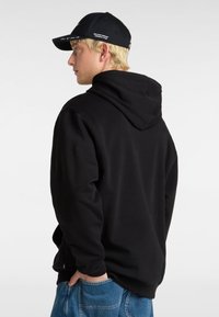 Felpa nera con cappuccio, dal texture morbido, con coulisse regolabile e tasca a marsupio; indossata con jeans in denim larghi e un cappellino nero.