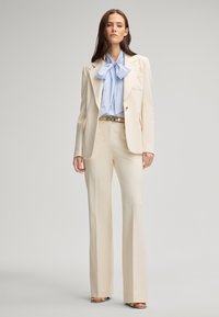 Cream-colored tailored blazer with matching wide-leg trousers. Features a light blue striped blouse with a large bow. Silver belt accent.
