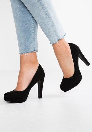 Czarne zamszowe szpilki z zaokrąglonym czubkiem, platformą i błyszczącym czarnym obcasem w kształcie szpilki, noszone z przetartymi jasnoniebieskimi dżinsami.