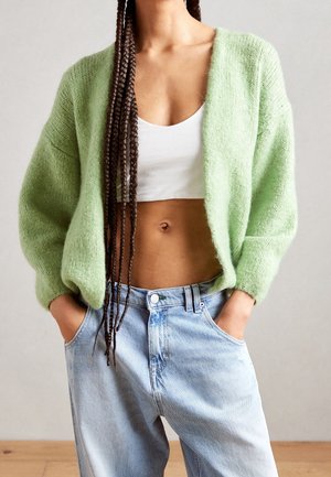 Young person with long braids wearing a light green cardigan, white crop top, and light blue high-waisted jeans with hands in pockets.