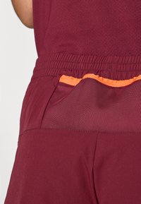 Bordové športové kraťasy s elastickým pásom, so žiarivo oranžovým akcentom na vrecku. Hladká textúra a priedušný materiál.