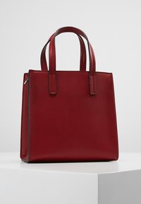 Punainen nahkainen käsilaukku, jossa on sileä pinta, neliömäinen muoto ja kaksi lyhyttä kahvaa. Sisältää kontrastivärisiä tikkejä ja vetoketjuyksityiskohdan yhdellä sivulla.