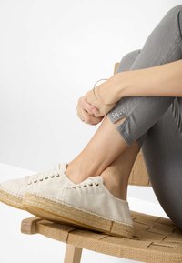 Lichtgrijze slim fit broek met uitsnijdingen en versieringen aan de zoom. Witte canvas schoenen met een geweven espartosole. Houten stoel.