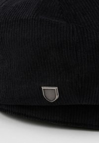 Fekete bordás sapka, texturált felülettel, ívelt formával és egy ezüst pajzs logóval az oldalon.