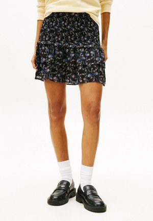 Femme portant une mini-jupe noire à fleurs, des chaussettes blanches, des mocassins noirs et un haut jaune clair à manches longues, debout les mains dans les poches.