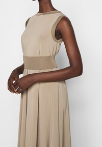 Robe sans manches beige avec un col et une taille côtelés, présentant une texture lisse et un design de jupe plissée.
