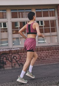 Sport bra in dark pink and black with a racerback design, paired with loose dark pink shorts. Wears green sneakers with white soles and socks.