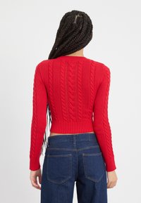 Rode, ribbelgebreide trui met kabelpatronen. Aansluitende, cropped lengte en lange mouwen. Gecombineerd met donkerblauwe jeans.