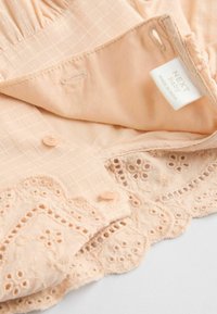 Tissu en coton de couleur pêche avec un motif à carreaux, comportant une garniture en dentelle décorative et des boutons. L'étiquette indique « NEXT baby, fabriqué en Inde. »