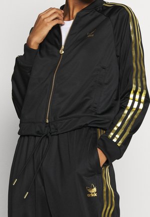 Person iført sort Adidas tracksuit med guldstriber og logoer, hånd justerer lynlåsen på jakken.