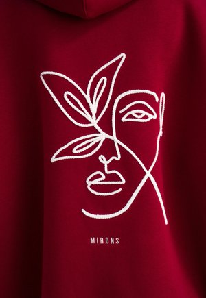 Felpa rossa con cappuccio, decorata con un design ricamato di un volto bianco e accenti di foglia, con la scritta "MIRONS" sotto il grafico. Texture del tessuto liscia.