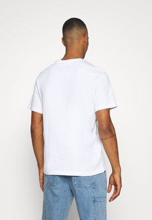 Homme portant un t-shirt blanc uni à manches courtes et un jean bleu clair, debout dos à la caméra sur un fond blanc.