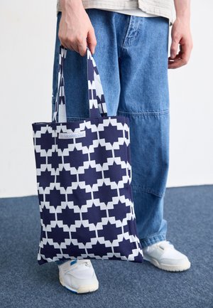 Marinefarbener Einkaufsbeutel mit einem geometrischen weißen Muster, aus robustem Stoff gefertigt, mit zwei Griffen und einem Etikett mit der Aufschrift "On Vacation Club."