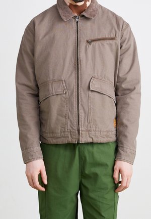 Mężczyzna w brązowej kurtce na zamek z kołnierzem z sztruksu i zielonych spodniach, stojący na jednolitym białym tle.