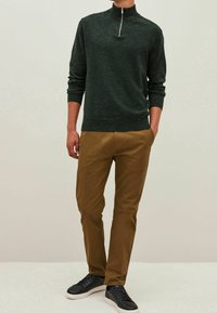 Tmavě zelený pulovr s vysokým límcem a polo zipem, ve spojení s béžovými džínami. Model má na sobě černé tenisky. Hladké textury, neformální design.