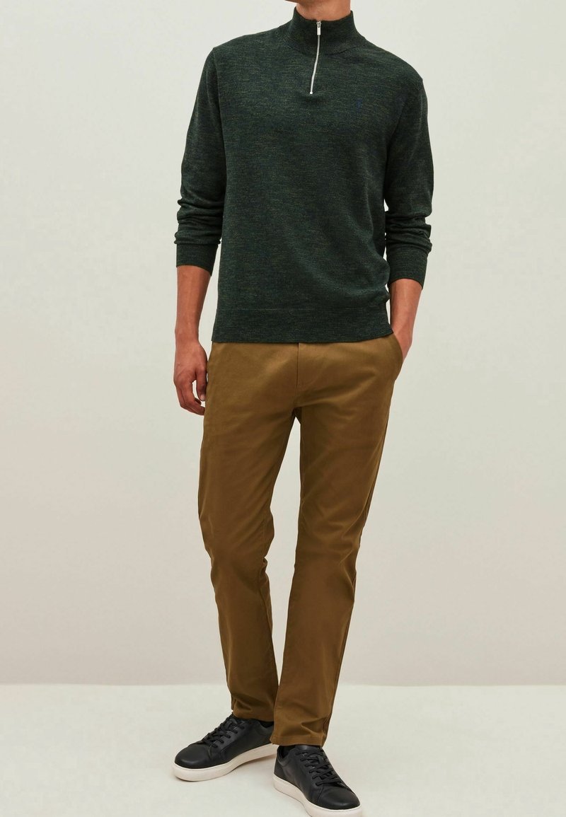 Tmavě zelený pulovr s vysokým límcem a polo zipem, ve spojení s béžovými džínami. Model má na sobě černé tenisky. Hladké textury, neformální design.