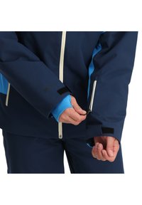 Marineblaue wasserdichte Jacke mit hellblauen Akzenten, Reißverschlussmanschetten und Klettverschlüssen. Verfügt über einen kontrastierenden weißen Reißverschluss und ein Logo auf dem Ärmel.