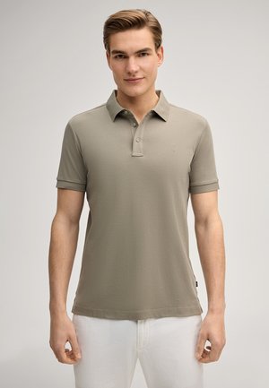 Giovane uomo che indossa una polo beige a maniche corte e pantaloni bianchi, in piedi davanti a uno sfondo chiaro e semplice.