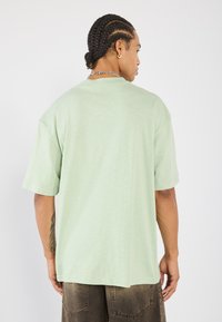 Světlonzelené oversized tričko s kulatým výstřihem a krátkými rukávy, vyrobené z texturované látky. Zadní pohled ukazuje jednoduchost designu.