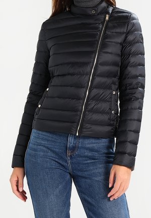 Woman wearing a black quilted jacket with an asymmetrical front zipper, paired with blue jeans and a gray turtleneck sweater.